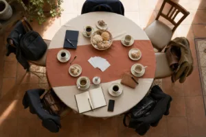 Mesa redonda de desayuno de negocios vista desde arriba, con tazas de café, pan dulce y tarjetas de presentación; una silla vacía